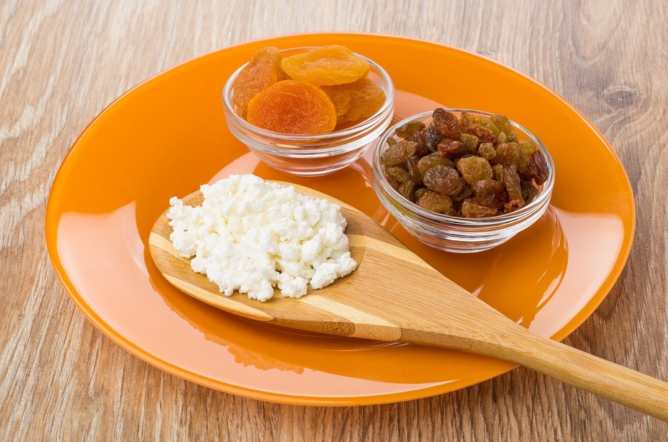 Bamboo spoon with grainy cottage cheese, bowls with raisins and dried apricots in orange plate on wooden table