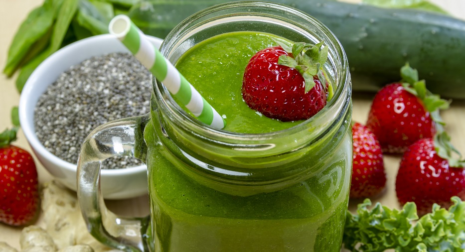 Healthy green juice smoothie surrounded by whole fruits, vegetables and chia seeds with fresh strawberry garnish and green swirl straw